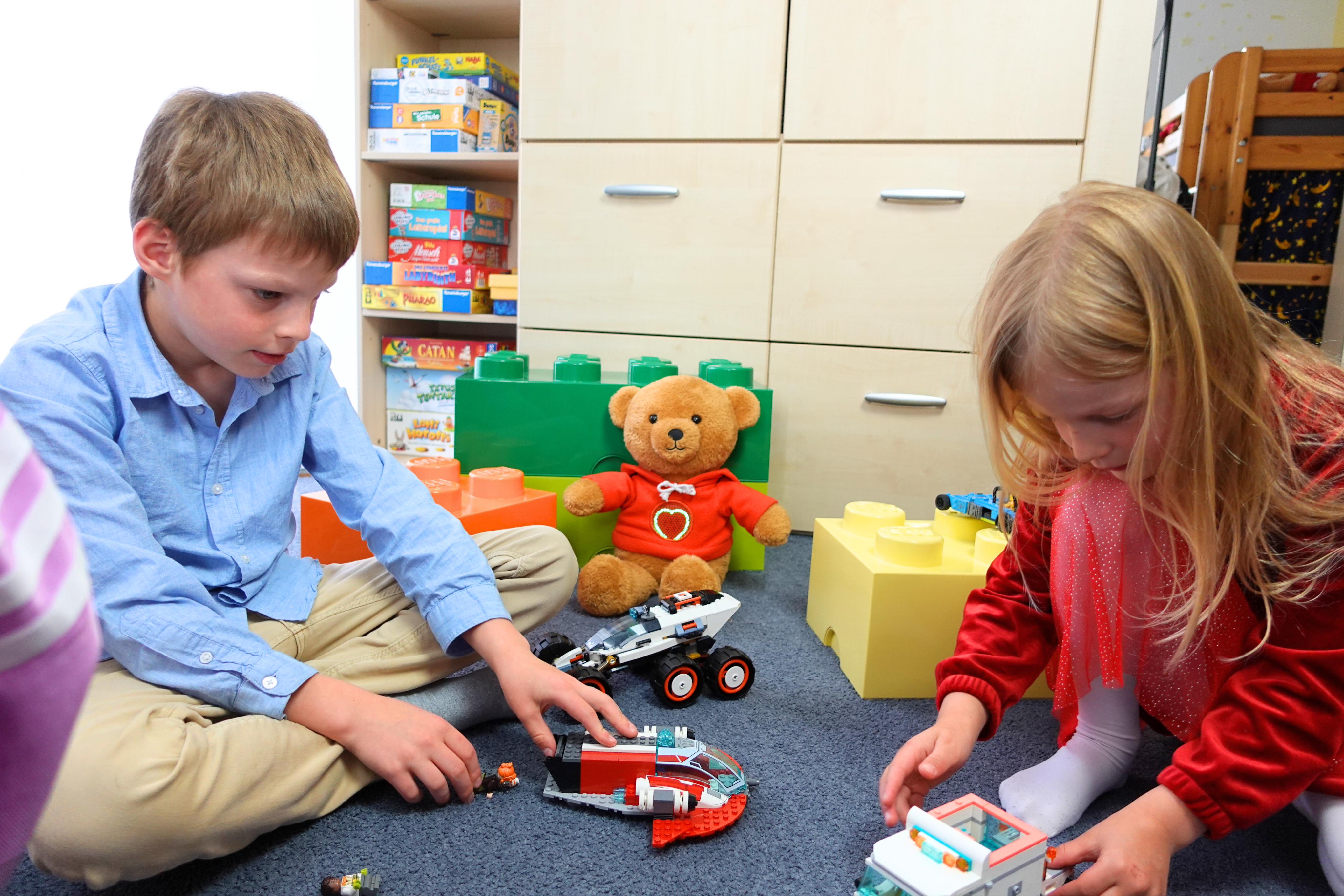 Ein Junge und ein Mädchen sitzen auf dem Boden eines Kinderzimmers und spielen mit Bausteinen und Fahrzeugen. Im Hintergrund sitzt der MyMilo Teddybär mit einem grün leuchtenden Herzen auf seinem Pullover. Beide Kinder wirken konzentriert auf ihr Spiel vertieft.