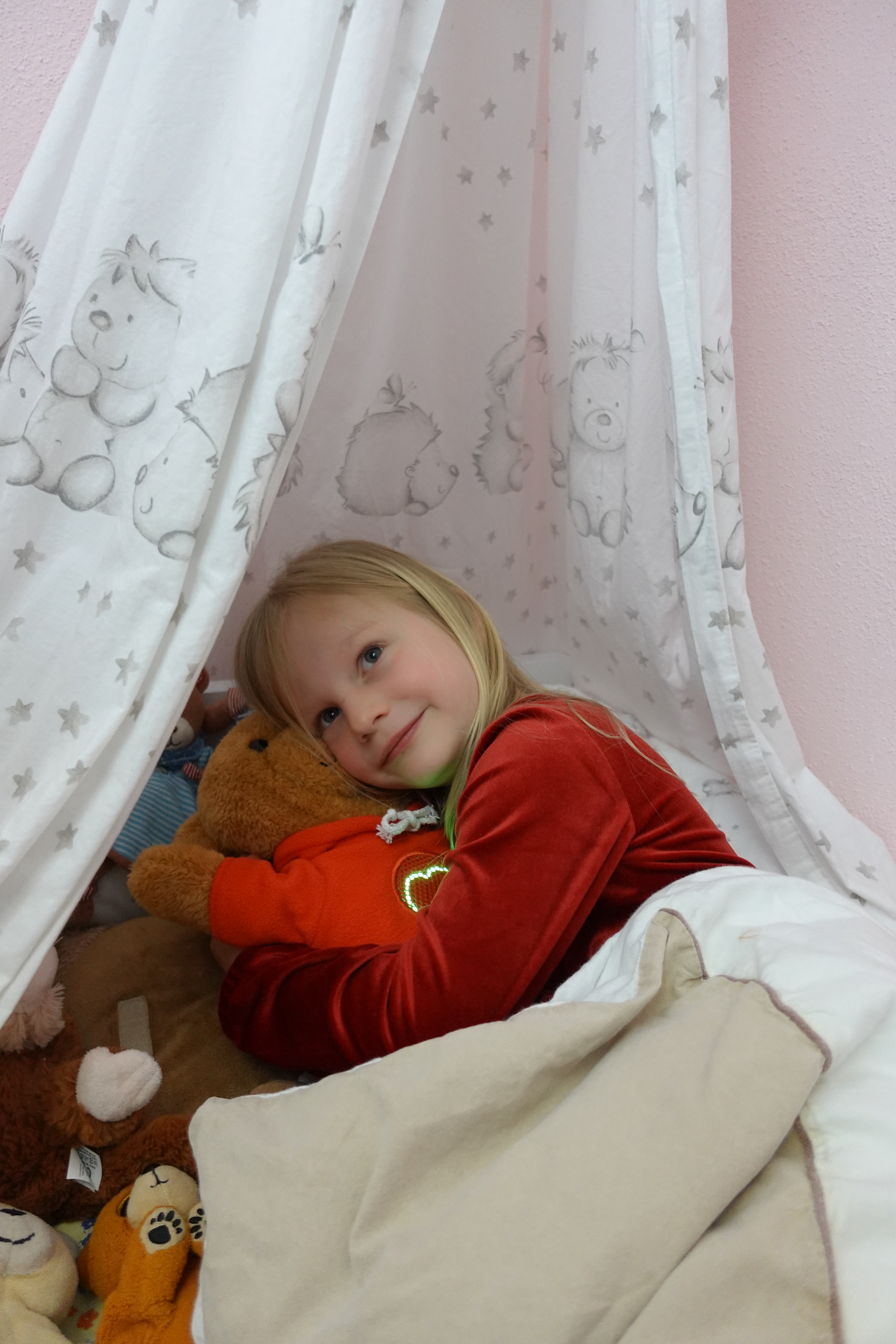 Ein Mädchen liegt gemütlich unter einem weißen Baldachin mit Sternen- und Tiermotiven und kuschelt sich an einen MyMilo Teddybär mit leuchtendem Herzsymbol auf seinem Pullover. Sie wirkt entspannt und zufrieden und schaut verträumt in ihr Zimmer. Um sie herum liegen weitere Stofftiere.