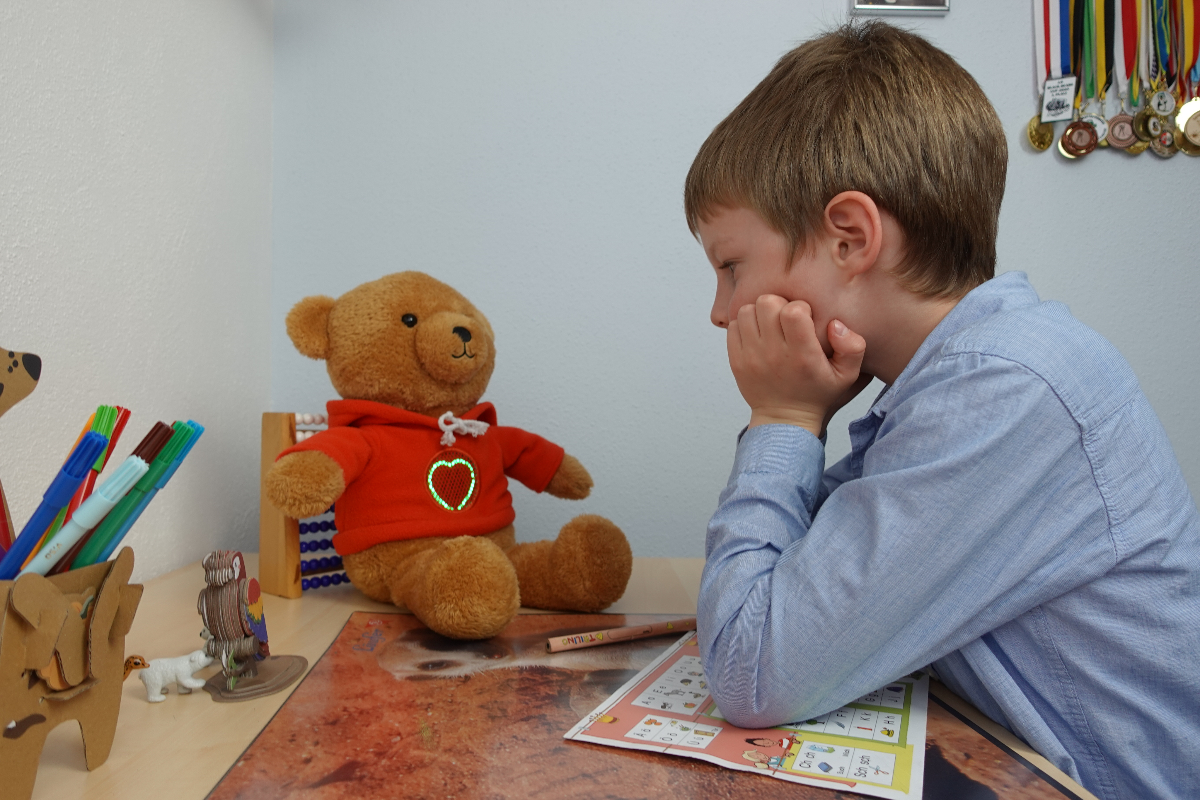 Ein Junge sitzt an einem Schreibtisch und arbeitet konzentriert mit einem Bleistift an einem Übungsblatt.  Ein MyMilo Teddybär mit leuchtendem Herzen auf seinem Pullover sitzt ihm gegenüber. Im Hintergrund sind einige Medaillen an der Wand zu sehen.
