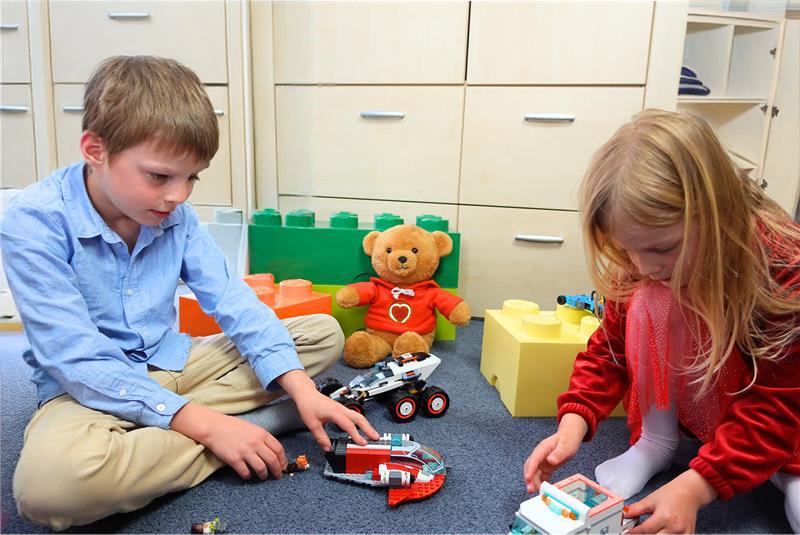 Ein Junge und ein Mädchen sitzen auf dem Boden eines Kinderzimmers und spielen mit Bausteinen und Fahrzeugen. Im Hintergrund sitzt der MyMilo Teddybär mit einem grün leuchtenden Herzen auf seinem Pullover. Beide Kinder wirken konzentriert auf ihr Spiel vertieft.