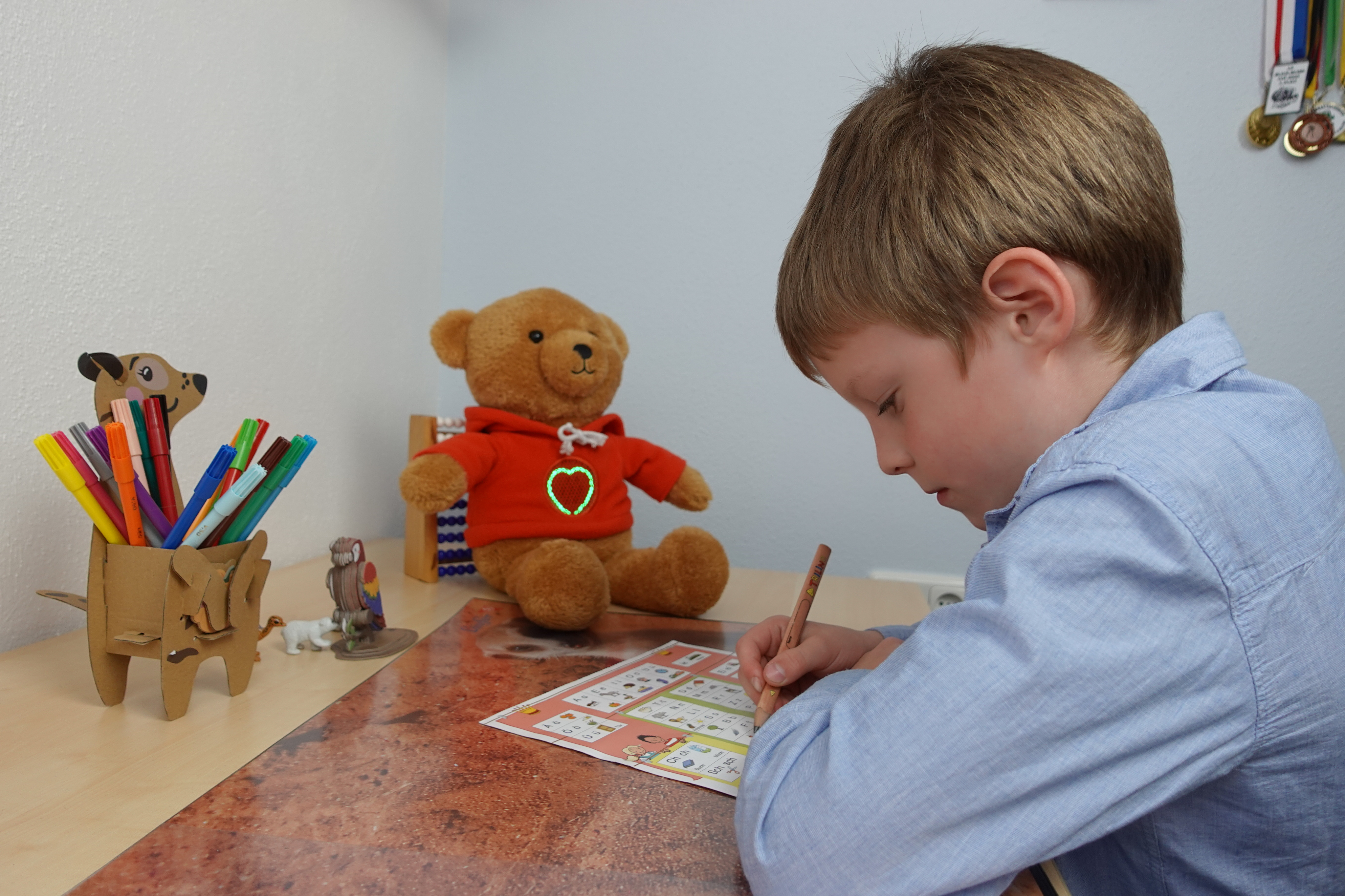 Ein Junge sitzt an einem Schreibtisch und arbeitet konzentriert mit einem Bleistift an einem Übungsblatt.  Ein MyMilo Teddybär mit leuchtendem Herzen auf seinem Pullover sitzt ihm gegenüber. Im Hintergrund sind einige Medaillen an der Wand zu sehen.